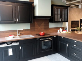Winkelküche im modernen Landhausstil mit schwarzer Lackfront, 2 Hängeschränken, davon einer mit Glastüren. Arbeitsplatte und Nischenverkleidung in einen warmen Braunton (=Ponderosa Pine Nachbildung). Die Blancospüle ist im hellen grau.
